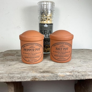 May include: A terracotta pepper pot and salt pot with matching lids, and a clear glass grinder filled with sea salt. The pepper pot and salt pot have labels with text. The items are on a rustic wooden surface.