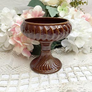 May include: A brown ceramic pedestal bowl with a fluted design. The bowl is on a white lace tablecloth.