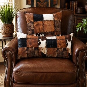 Può includere: Due cuscini decorativi con un design patchwork composto da pannelli testurizzati marroni, neri e bianchi. Il cuscino quadrato è posizionato dietro un cuscino rettangolare, entrambi realizzati con cuciture scure visibili tra i riquadri.
