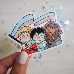 May include: A sticker featuring three cartoon characters with determined expressions. One character holds a transgender pride flag. The sticker has a white border and is held by a hand. Glittery confetti is scattered around the sticker.