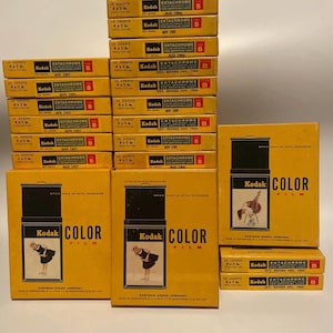 May include: A stack of vintage Kodak film boxes, predominantly yellow with black and red text. The boxes are labelled "Ektachrome" and "Color Film," with dates from the 1960s. Some boxes feature a woman in a dress.