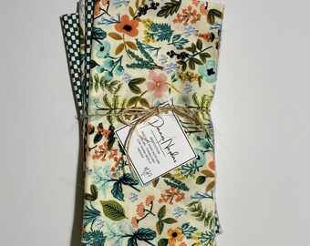Könnte beinhalten: Ein Stapel von vier Stoffservietten mit einem Blumenmuster in Grün-, Blau-, Orange- und Rosatönen. Die Servietten sind mit Schnur zusammengebunden und mit einem kleinen Etikett versehen, auf dem "Donna Wolfe" steht.