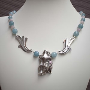 May include: A silver necklace with a unique pendant featuring a textured, organic shape. The pendant is accented with a small pink stone. The necklace is strung with light blue and pink beads.