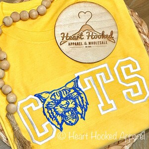 May include: Yellow t-shirt with a blue and white embroidered mascot and the letters "CTS" in white with a blue outline. The shirt is on a woven surface with a wooden tag that says "Heart Hooked Apparel & Wholesale Est. 2015".