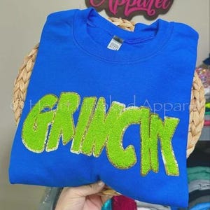 May include: A royal blue sweatshirt with the word "GRINCH" in lime green, outlined in gold. The sweatshirt is displayed on a woven basket. Above the sweatshirt is a wooden sign that says "Heart Hooked Apparel".