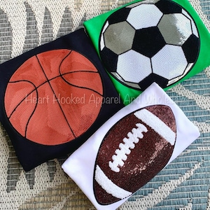 May include: Three t-shirts with different sports ball designs. The first shirt is black with a brown basketball, the second shirt is green with a white and black soccer ball, and the third shirt is white with a brown football.
