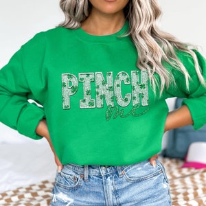 May include: Green sweatshirt with the text "PINCH ME" in a glittery, textured design. The word "PINCH" features shamrock accents. The sweatshirt is a solid green color, perfect for St. Patrick's Day.