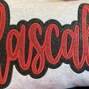 May include: A gray sweatshirt with the word "Rascals" in a red glitter script font. The script is outlined in black with a glittery texture. The design is centered on the sweatshirt.