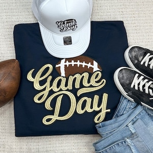 May include: A navy blue sweatshirt with a gold embroidered "Game Day" design and a brown football graphic. The sweatshirt is paired with a white trucker hat with the text "Game Day" and a pair of black sneakers.