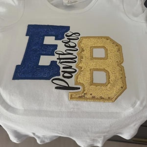 May include: A white t-shirt featuring a large blue sequined letter E on the left and a large gold sequined letter B on the right. Between the letters, the word Panthers is written in black cursive script, connecting the two sequined letters.