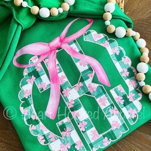 May include: Green and white plaid monogram with a pink bow. The monogram is surrounded by shamrocks and has a scalloped edge.