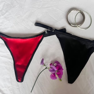 Peut inclure: Deux strings en satin, l'un rouge vif et l'autre noir profond, dotés de fines bretelles noires réglables avec des boucles en métal argenté. Deux anneaux circulaires en métal argenté sont placés au-dessus du string noir, et une petite branche de fleurs de pois de senteur violettes repose entre les deux vêtements.