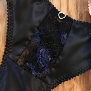 May include: Black satin lingerie with a lace panel featuring a floral design in shades of blue and black. The lace panel is bordered by a black satin trim. A small silver heart-shaped charm is attached to the top of the lace panel.