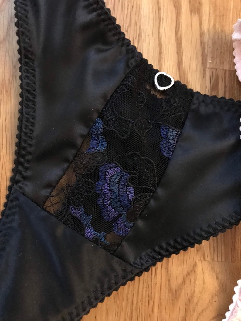 May include: Black satin lingerie with a lace panel featuring a floral design in shades of blue and black. The lace panel is bordered by a black satin trim. A small silver heart-shaped charm is attached to the top of the lace panel.