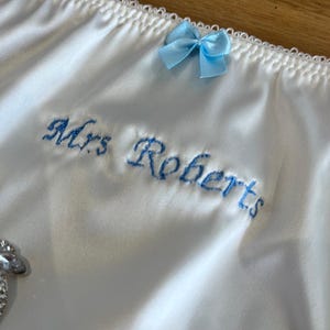 May include: White satin underwear with a delicate scalloped edge and a small light blue bow. The words "Mrs Roberts" are embroidered in light blue script. A decorative item is visible on the left.