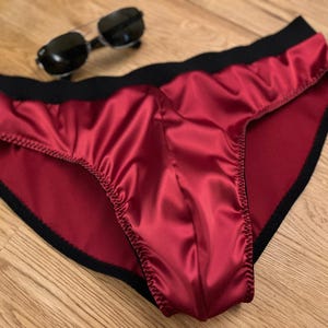 May include: Red satin underwear with black trim and waistband. Sunglasses and a silver wristwatch are also visible on a wooden surface.