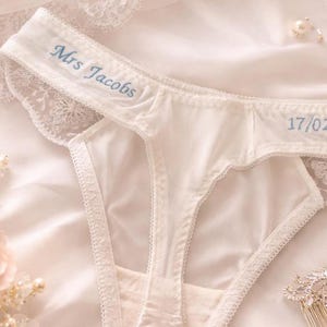 May include: White lace-trimmed thong featuring blue embroidered text that reads Mrs Jacobs on the left side and 17/02 on the right side. The garment is crafted from a smooth fabric with delicate floral lace edges along the waistband and leg openings.