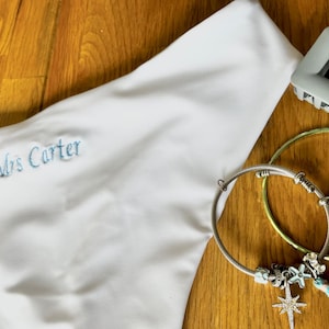 May include: A white garment bag embroidered with "Mrs Carter" in light blue. A silver charm bracelet with a turquoise tassel, a gold bracelet, and a silver hair clip are on a wooden surface.