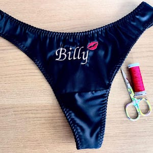 May include: Navy blue satin thong underwear with the name "Billy" embroidered in white, along with a red lip design. A spool of red thread and decorative scissors are next to the underwear.