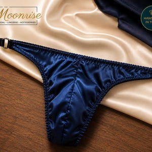 Peut inclure: Un string en satin bleu foncé avec un design en tissu froncé et des anneaux métalliques argentés de chaque côté. Le vêtement porte le texte Moonrise Bridal Lingerie Accessories et un badge circulaire indiquant Handmade in the UK.