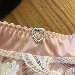 May include: A close-up of a pink satin and lace garment with a silver heart-shaped button.