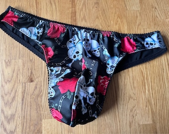 Op de afbeelding: Zwart ondergoed met een herhalend patroon van witte schedels, rode rozen en zilveren kettingen. Het kledingstuk heeft een gladde, satijnachtige afwerking met zwarte geschulpte elastische randen langs de tailleband en de beenopeningen.