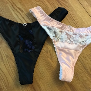 May include: Two pairs of satin thongs with lace trim. One thong is black with blue lace and the other is pink with white lace.