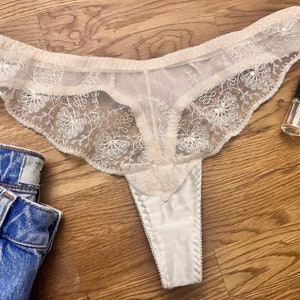May include: Ivory-colored thong underwear with delicate floral lace detailing. The lingerie features a satin front panel and scalloped edges. A pair of blue denim jeans and two bottles of nail polish are also in the image.