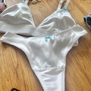 May include: White satin lingerie set with delicate lace trim and light blue satin bows. The set includes a triangle bra and a thong.