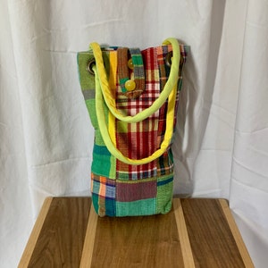 May include: A small, patchwork tote bag with a green, red, yellow, and blue plaid pattern. The bag has two yellow straps and a button closure.