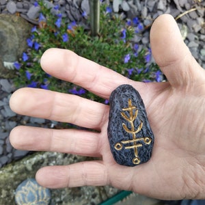 May include: A black stone with a gold tree-like symbol carved into it. The stone is being held in a hand.