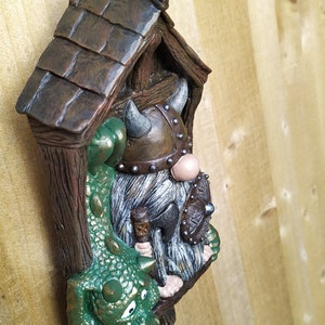 K&ouml;nnte beinhalten: Detaillierte Figur eines Wikinger-Zwergs mit einem Drachen. Der Zwerg tr&auml;gt einen geh&ouml;rnten Helm und h&auml;lt eine kleine Axt und einen Schild. Der Drache ist gr&uuml;n mit goldenen Akzenten. Die Figur befindet sich in einer Holzstruktur.
