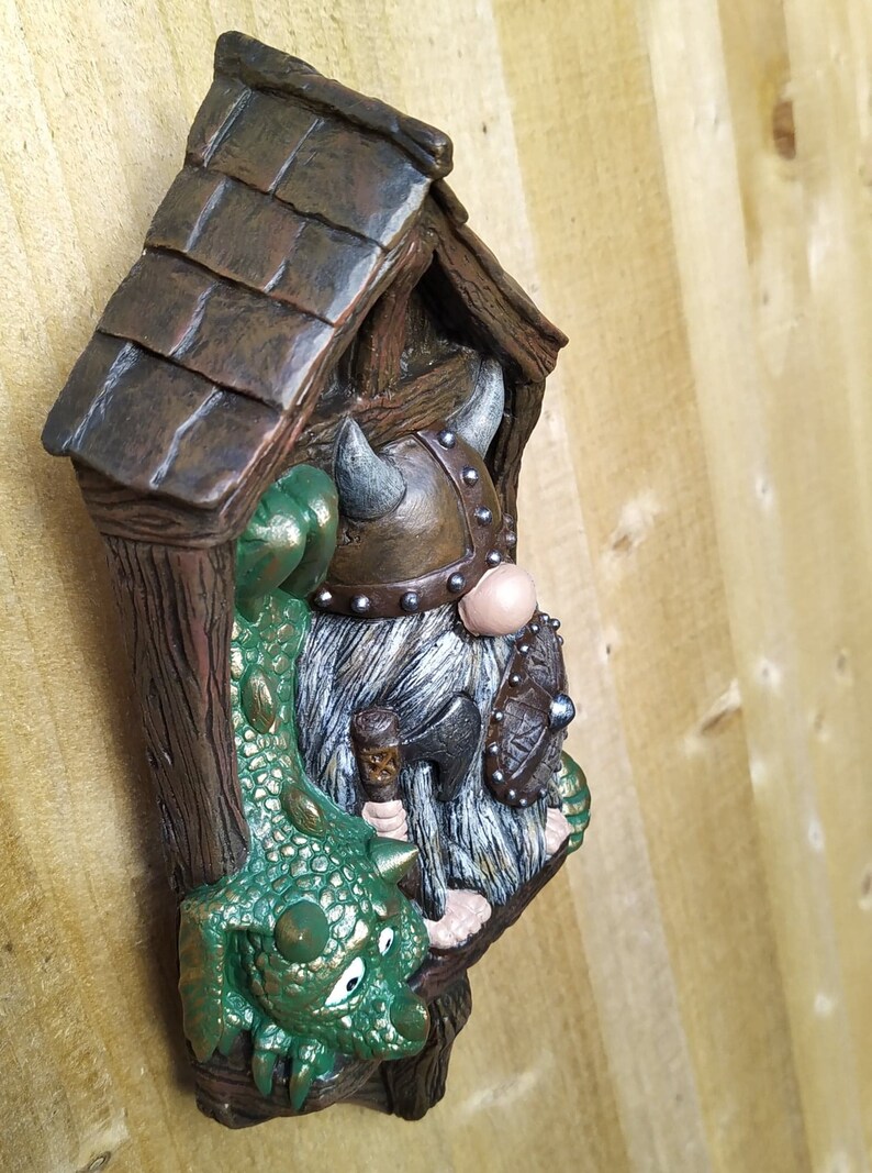 K&ouml;nnte beinhalten: Detaillierte Figur eines Wikinger-Zwergs mit einem Drachen. Der Zwerg tr&auml;gt einen geh&ouml;rnten Helm und h&auml;lt eine kleine Axt und einen Schild. Der Drache ist gr&uuml;n mit goldenen Akzenten. Die Figur befindet sich in einer Holzstruktur.