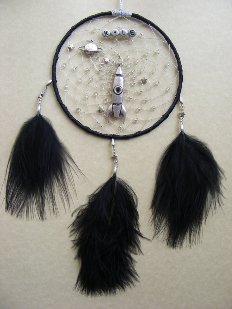 Pu&ograve; includere: Acchiappasogni nero con piume nere, perline argentate e un ciondolo a razzo argentato. L'acchiappasogni ha il nome "KYLE" scritto in perline argentate.