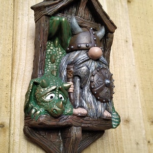 K&ouml;nnte beinhalten: Skurrile Wanddekoration mit einem Wikinger-Zwerg und einem gr&uuml;nen Drachen. Der Zwerg tr&auml;gt einen geh&ouml;rnten Helm und h&auml;lt Schild und Axt. Der Drache hat einen traurigen Gesichtsausdruck. Das St&uuml;ck besteht aus braunem, strukturiertem Material.