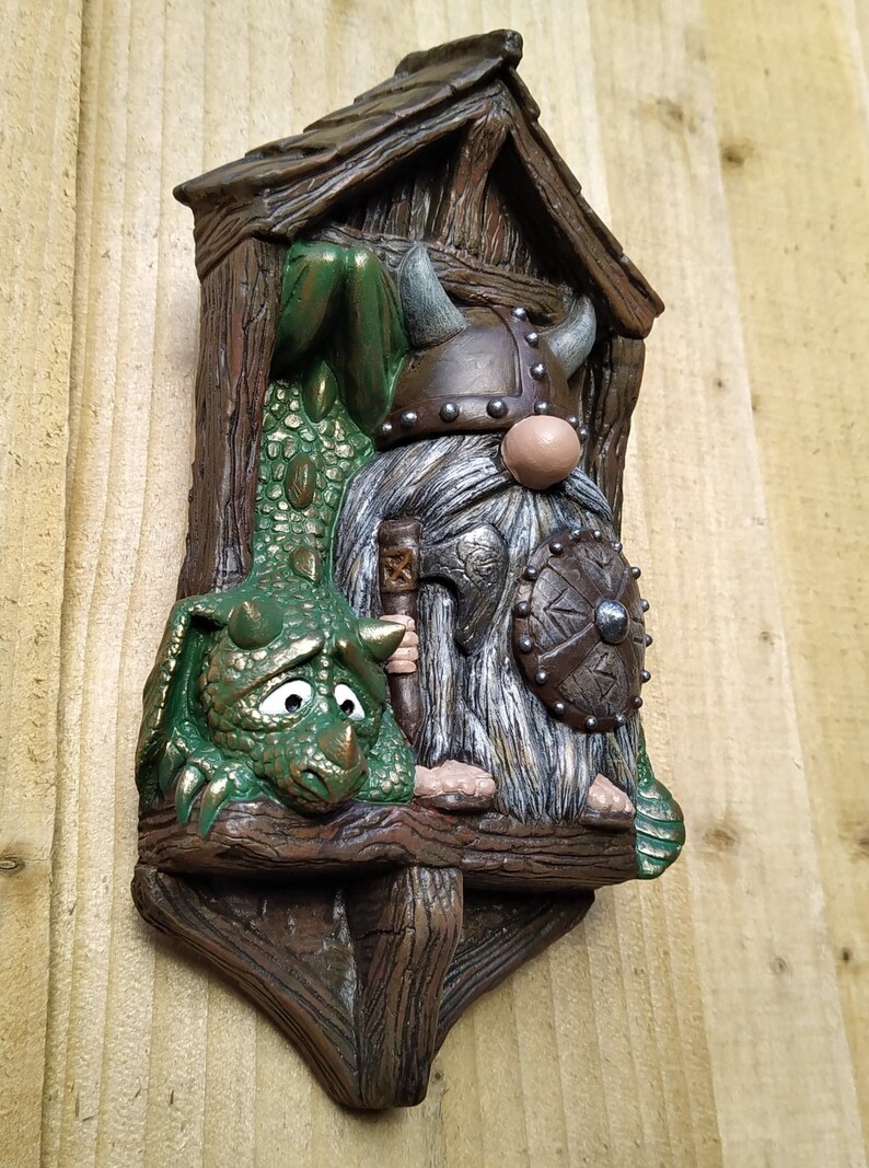 K&ouml;nnte beinhalten: Skurrile Wanddekoration mit einem Wikinger-Zwerg und einem gr&uuml;nen Drachen. Der Zwerg tr&auml;gt einen geh&ouml;rnten Helm und h&auml;lt Schild und Axt. Der Drache hat einen traurigen Gesichtsausdruck. Das St&uuml;ck besteht aus braunem, strukturiertem Material.