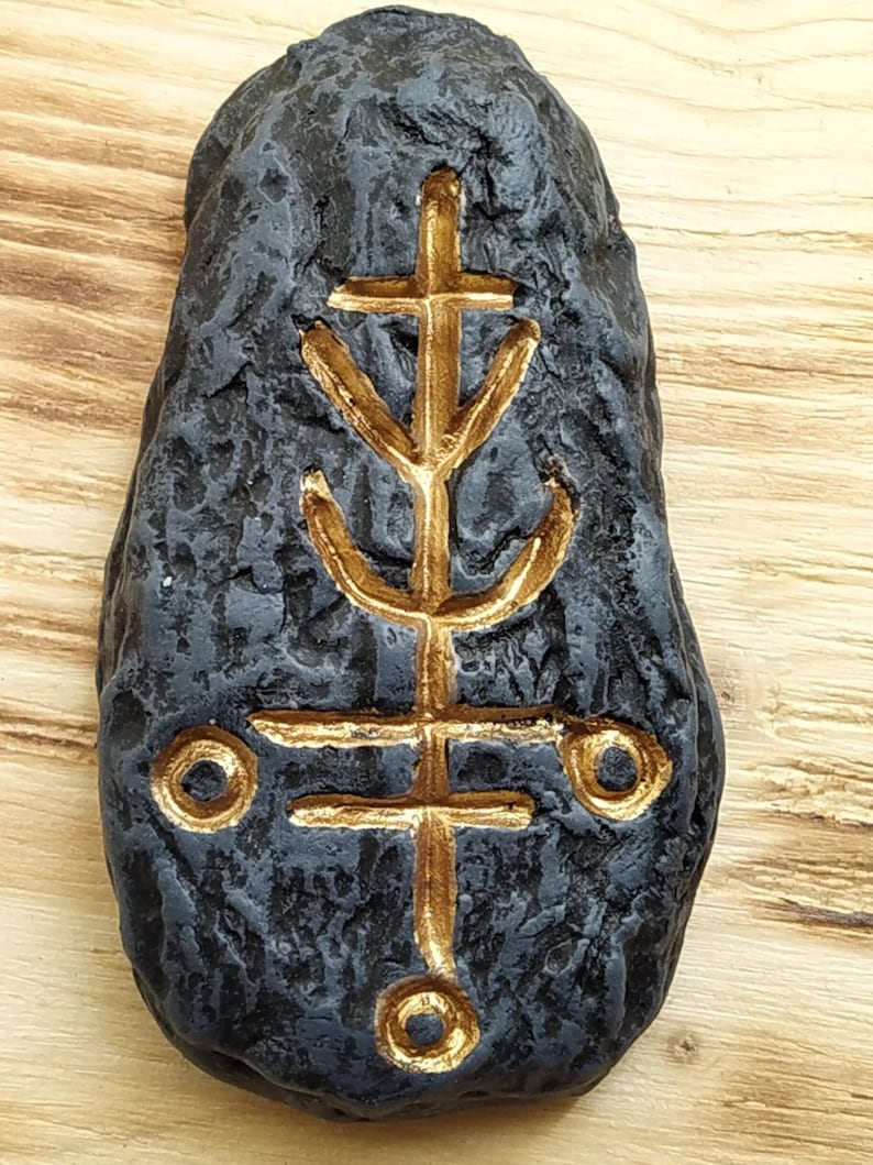 May include: A black stone with a gold symbol carved into it. The symbol is a cross with a Y shape at the bottom and two circles on either side.