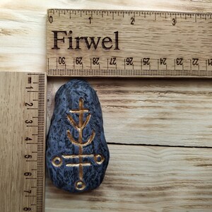 May include: A black stone with a gold symbol carved into it. The symbol is a stylized tree with a cross at the top. The stone is about 2 inches tall and 1 inch wide. The word "Firwel" is written on a ruler in the background.