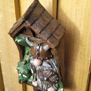 K&ouml;nnte beinhalten: Eine skurrile Gartenzwerg-Skulptur mit Wikingerhelm, Schild und einem gr&uuml;nen Drachen. Der Zwerg befindet sich in einer Miniatur-Holzstruktur mit strukturiertem Dach. Die Skulptur ist in Braun-, Gr&uuml;n- und Graut&ouml;nen bemalt.