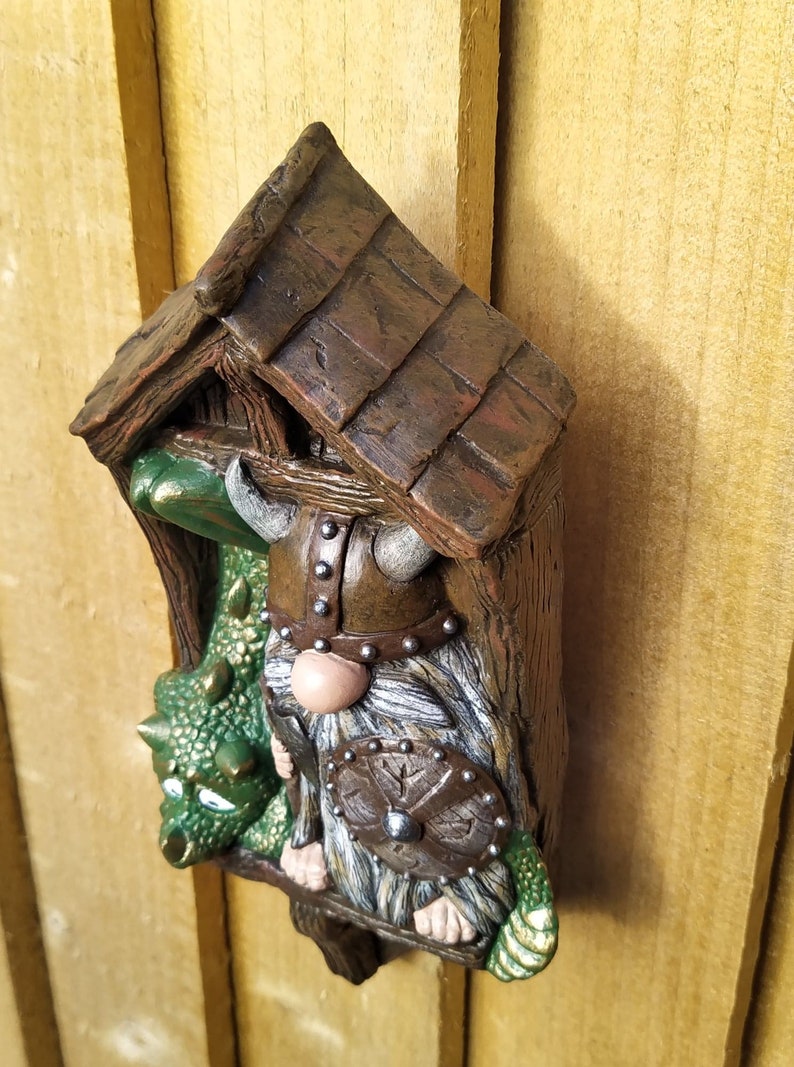 K&ouml;nnte beinhalten: Eine skurrile Gartenzwerg-Skulptur mit Wikingerhelm, Schild und einem gr&uuml;nen Drachen. Der Zwerg befindet sich in einer Miniatur-Holzstruktur mit strukturiertem Dach. Die Skulptur ist in Braun-, Gr&uuml;n- und Graut&ouml;nen bemalt.