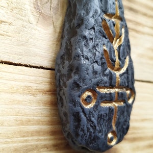 May include: A black stone with a gold design. The design is a stylized symbol with a cross shape and circles.