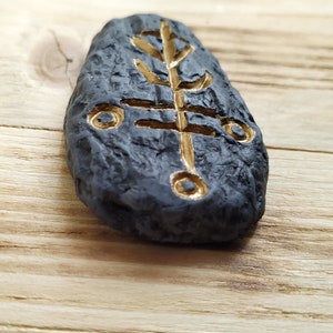 May include: A black stone with a gold tree symbol carved into it. The stone is smooth and has a textured surface.