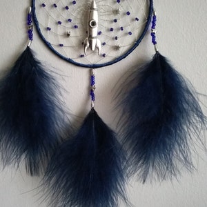 Pu&ograve; includere: Un acchiappasogni blu e argento con un razzo, un pianeta e stelle. L'acchiappasogni ha tre piume blu che pendono dal basso. L'acchiappasogni ha il nome "SAM" scritto su di esso.