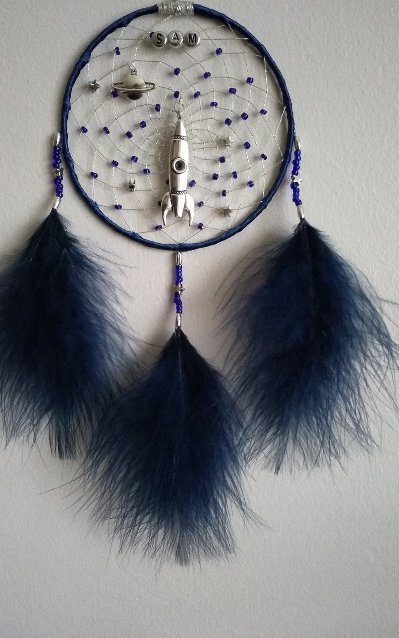 Pu&ograve; includere: Un acchiappasogni blu e argento con un razzo, un pianeta e stelle. L'acchiappasogni ha tre piume blu che pendono dal basso. L'acchiappasogni ha il nome "SAM" scritto su di esso.