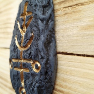 May include: A black stone with a gold-colored symbol etched into it. The stone is textured and has a rough surface. The symbol is a stylized design with a cross and a circle.