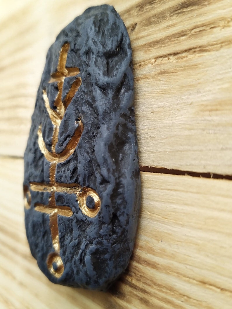 May include: A black stone with a gold-colored symbol etched into it. The stone is textured and has a rough surface. The symbol is a stylized design with a cross and a circle.