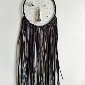 Pu&ograve; includere: Un acchiappasogni nero e argento con un razzo, pianeti e la parola "CAL" al centro. L'acchiappasogni ha frange nere che pendono dal basso.