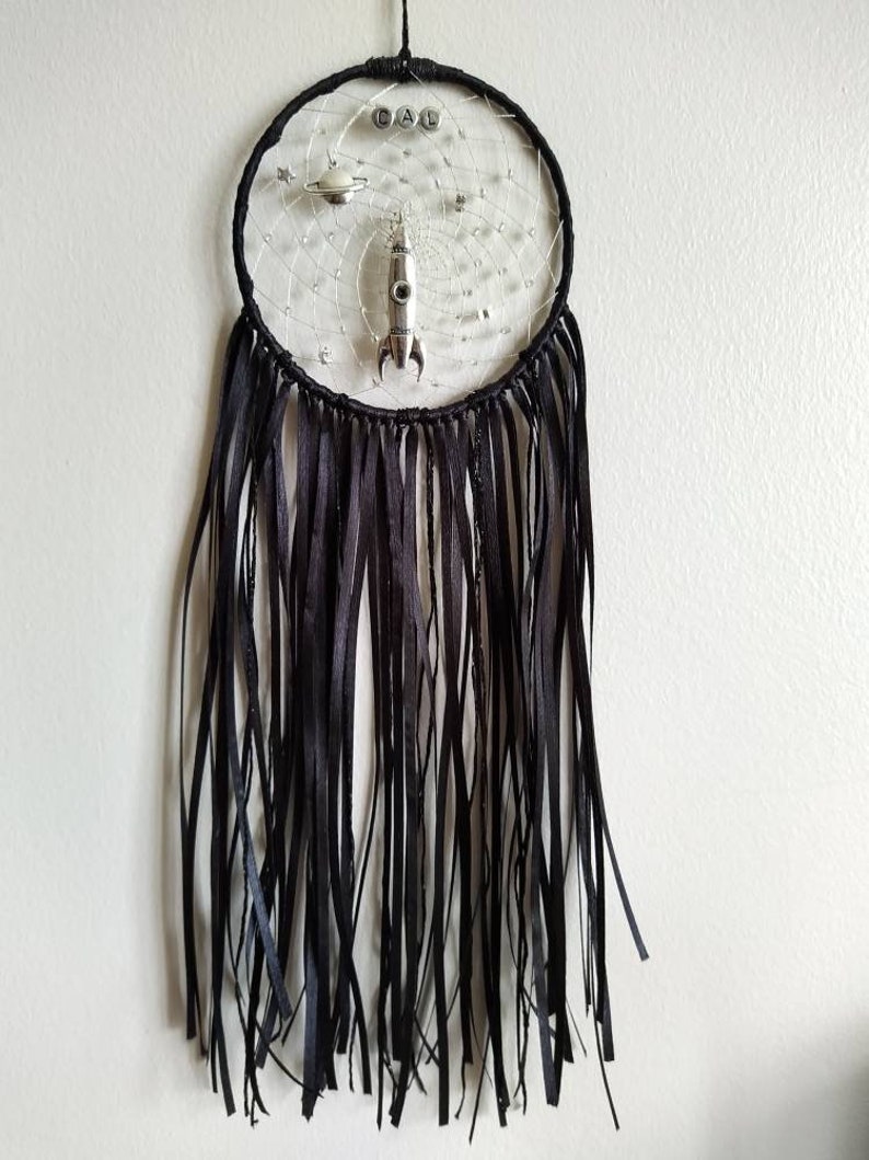 Pu&ograve; includere: Un acchiappasogni nero e argento con un razzo, pianeti e la parola "CAL" al centro. L'acchiappasogni ha frange nere che pendono dal basso.