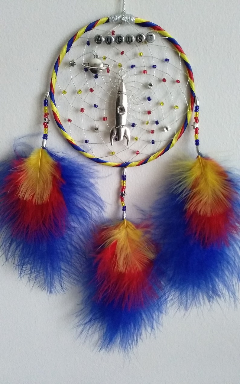 Pu&ograve; includere: Un acchiappasogni giallo, rosso e blu con la parola "AUGUST" scritta in perline d'argento. L'acchiappasogni ha un razzo d'argento e un ciondolo di pianeta d'argento che pendono. Tre piume multicolori pendono dalla parte inferiore dell'acchiappasogni.