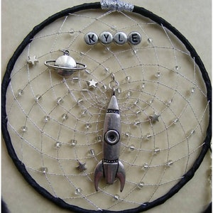 Pu&ograve; includere: Un acchiappasogni nero con una rete di perline argentate, che presenta un razzo argentato, un pianeta argentato con anelli e perline argentate che compongono il nome "KYLE".